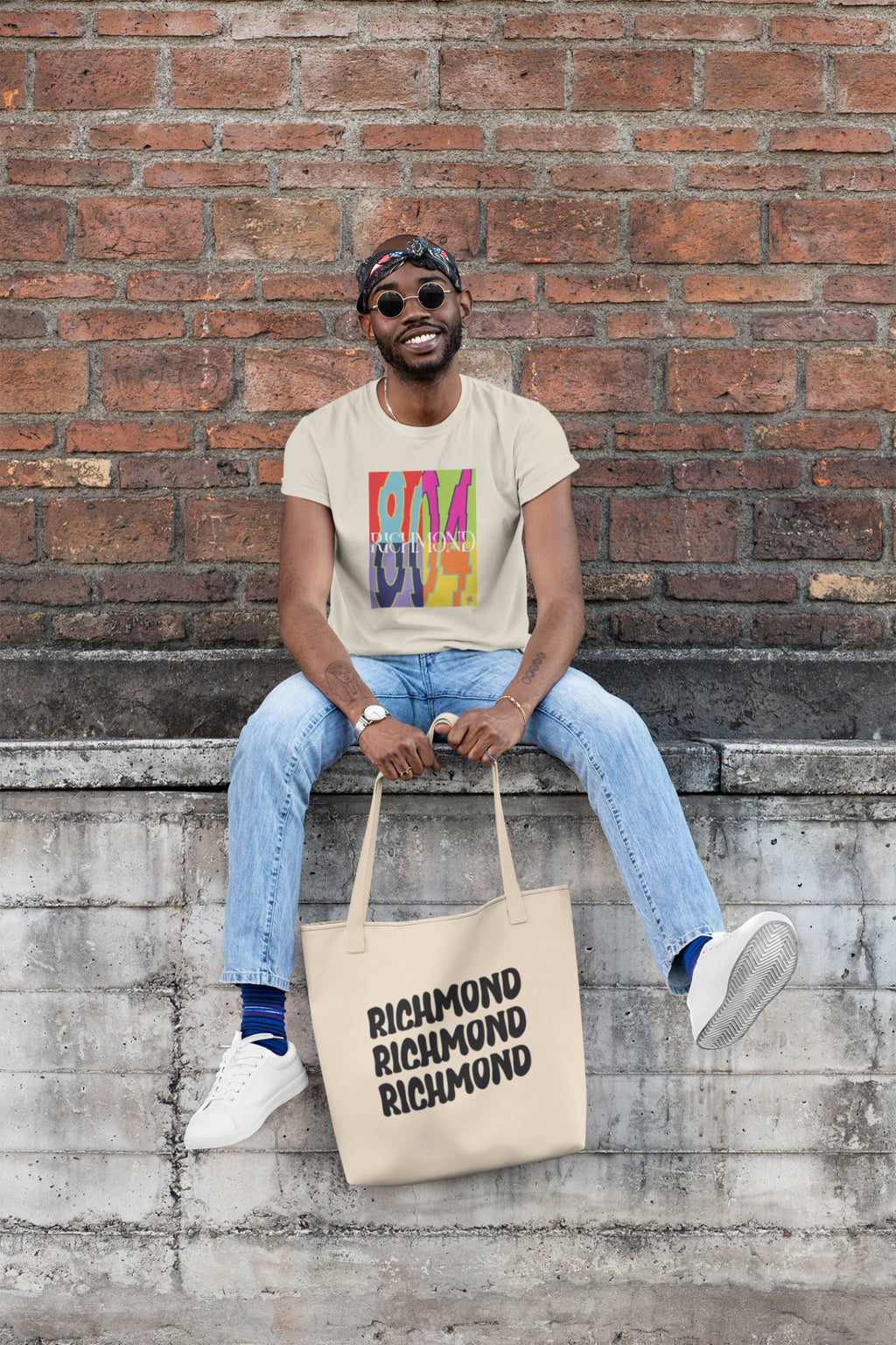 Man sitting on a brick wall, holding a tote bag wearing the new Already Richmond 804 Pop-Art Tee Shirt.
