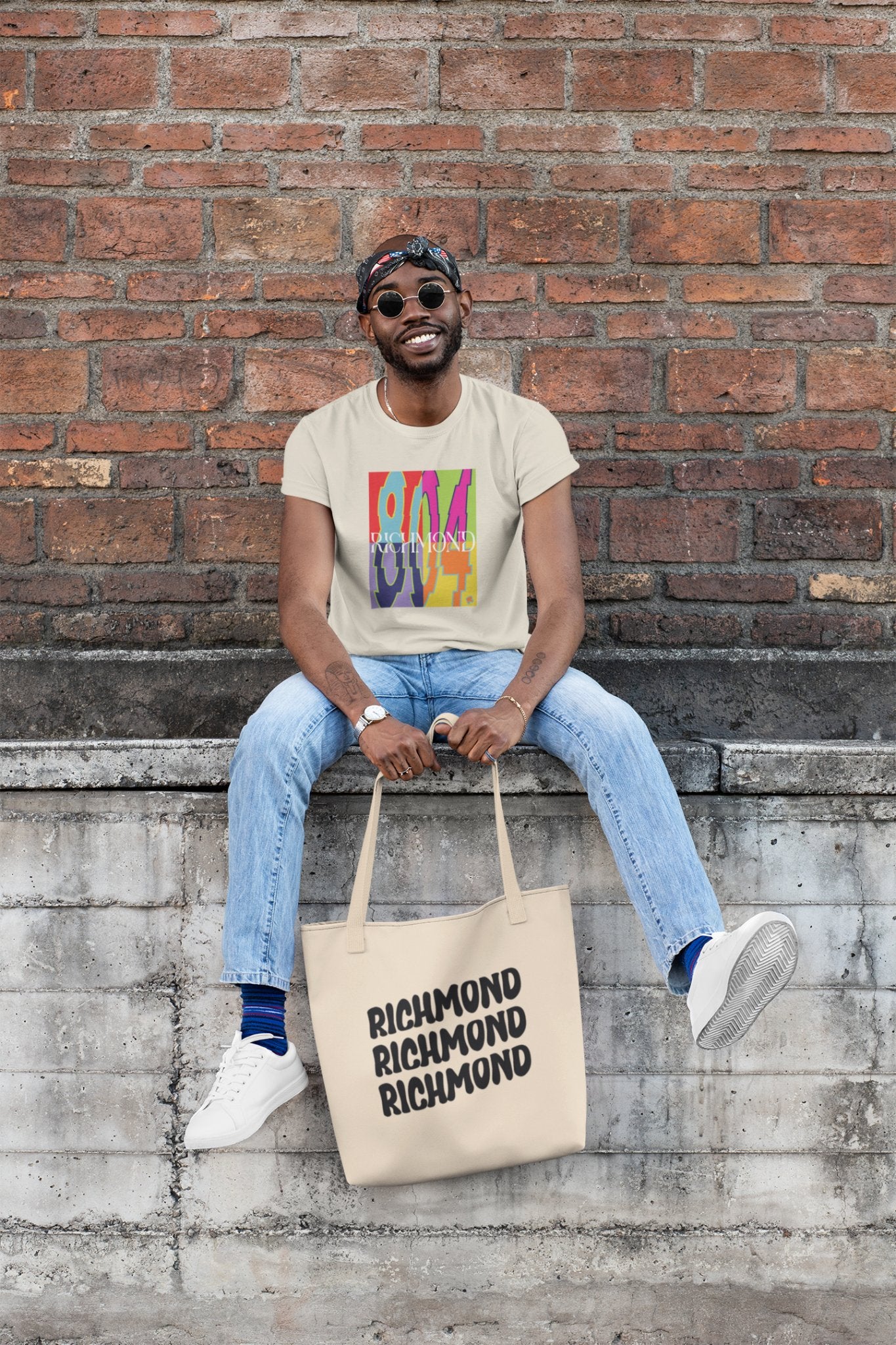 Man sitting on a brick wall, holding a tote bag wearing the new Already Richmond 804 Pop-Art Tee Shirt.